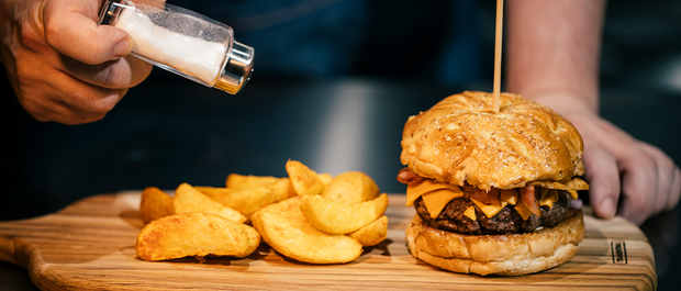 Cheeseburger with bacon and potato wedges being seasoned on a wooden board