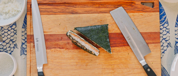Two triangular sushi sandwiches with salmon and rice on a wooden board beside kitchen knives